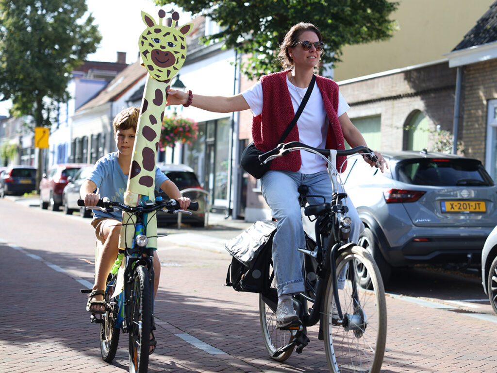 jongen op een fiets waarbij aan het stuur een hoofd van een giraffe is bevestigd - fietsend naast zijn moeder.