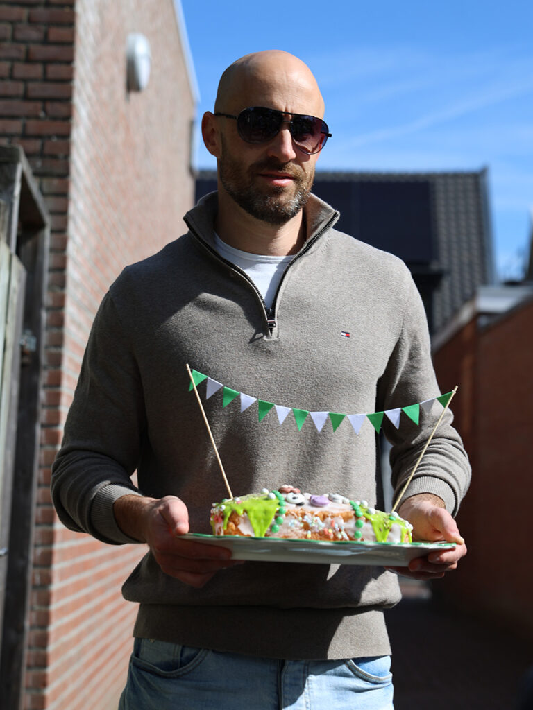 Vader komt aangelopen en houdt een bord vast met daarop een taart met groene en witte vlaggetjes
